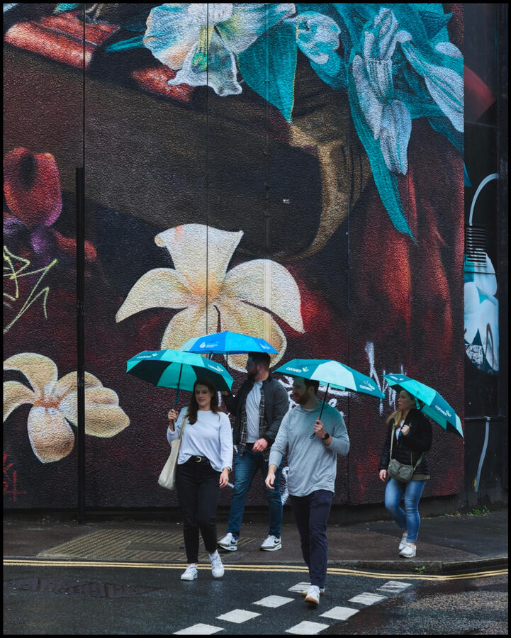 Blue Monday People crossing road with bright blue umbrellas with large blue mural on wall behind them